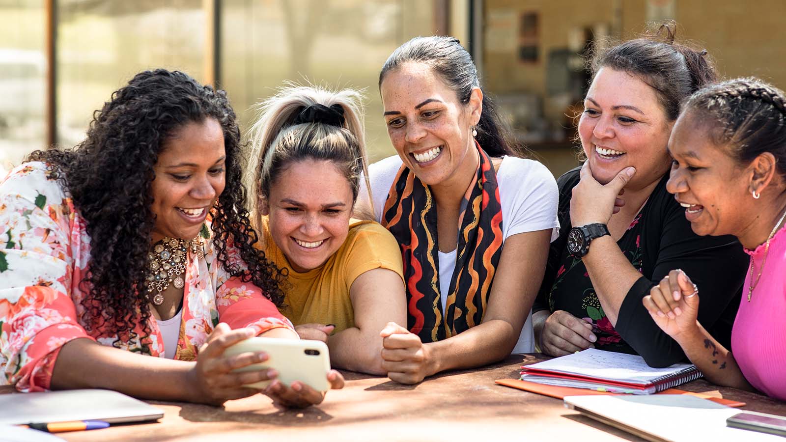 a group of five diverse women sitting together laughing together looking at phone