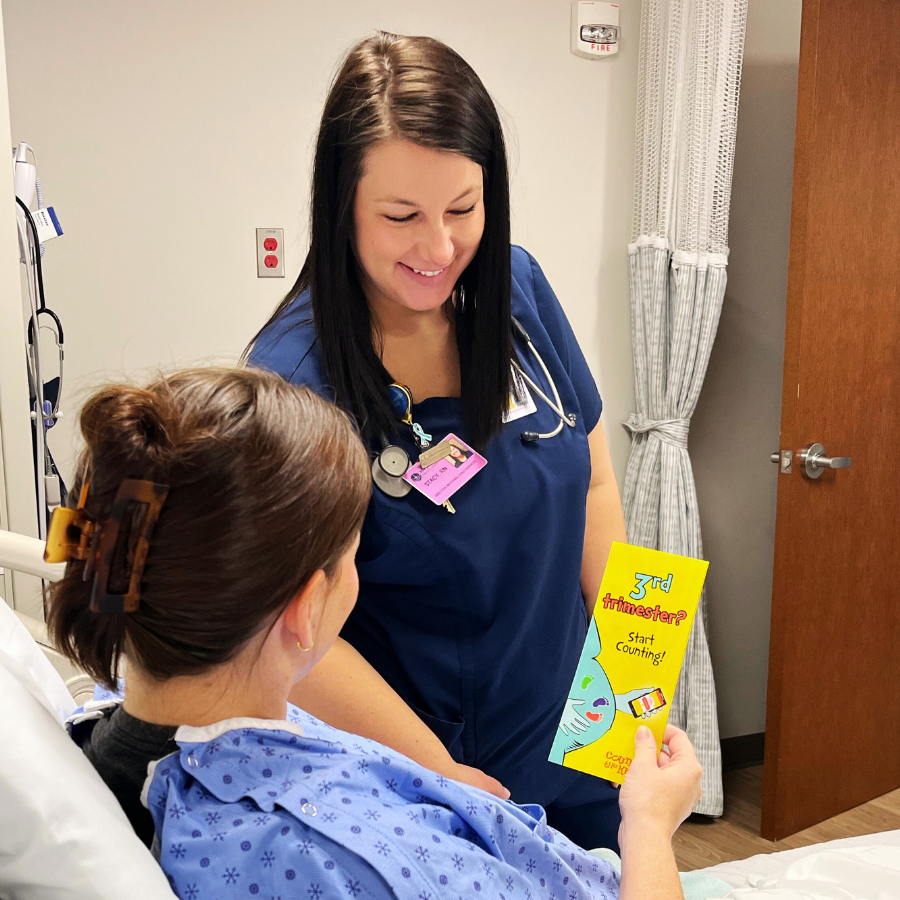 A nurse in blue scrubs and a patient in a hospital gown share smiles in a hospital room. The patient holds a Count the Kicks brochure. The mood is caring and supportive.