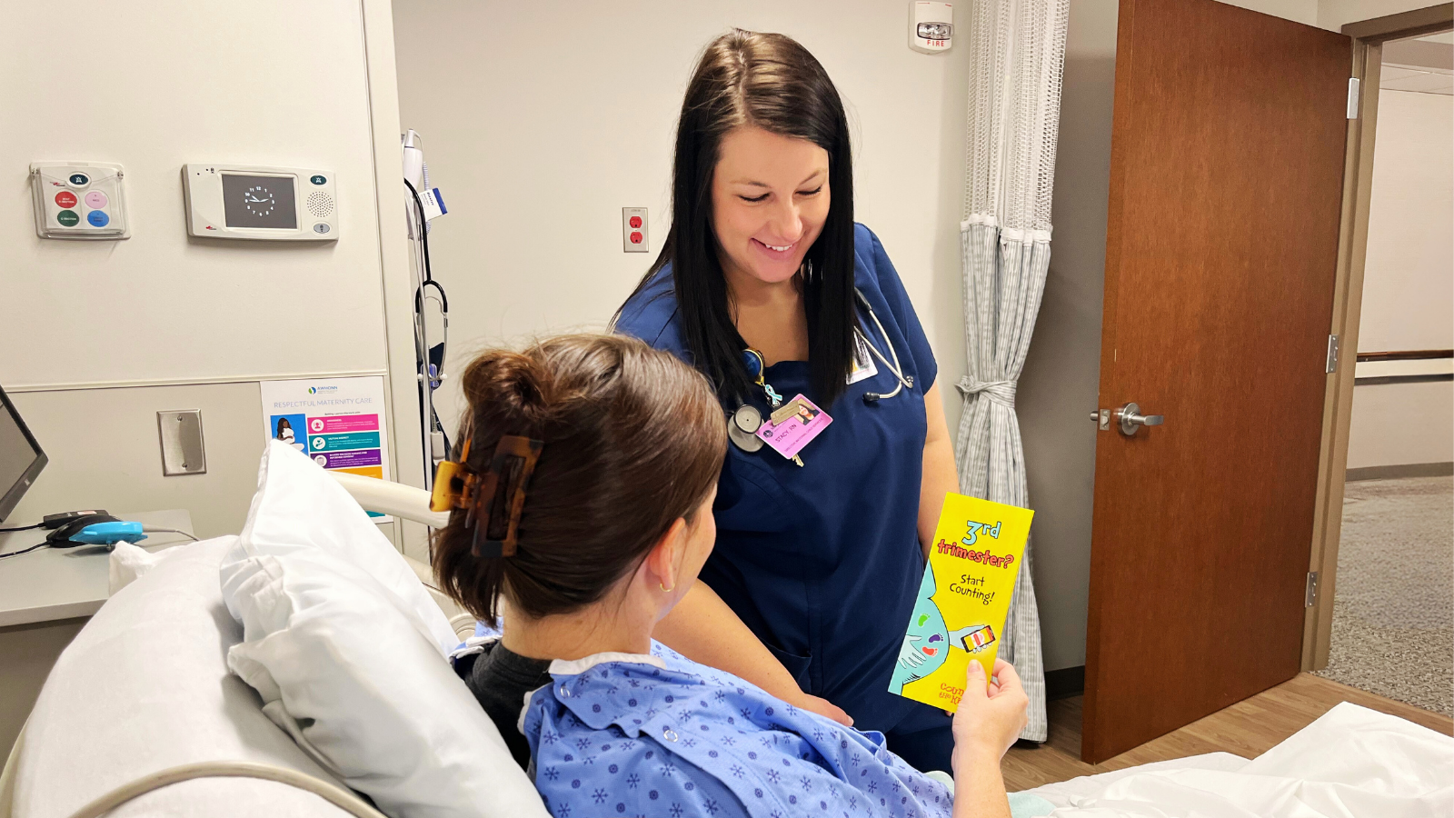 A nurse in blue scrubs and a patient in a hospital gown share smiles in a hospital room. The patient holds a Count the Kicks brochure. The mood is caring and supportive.