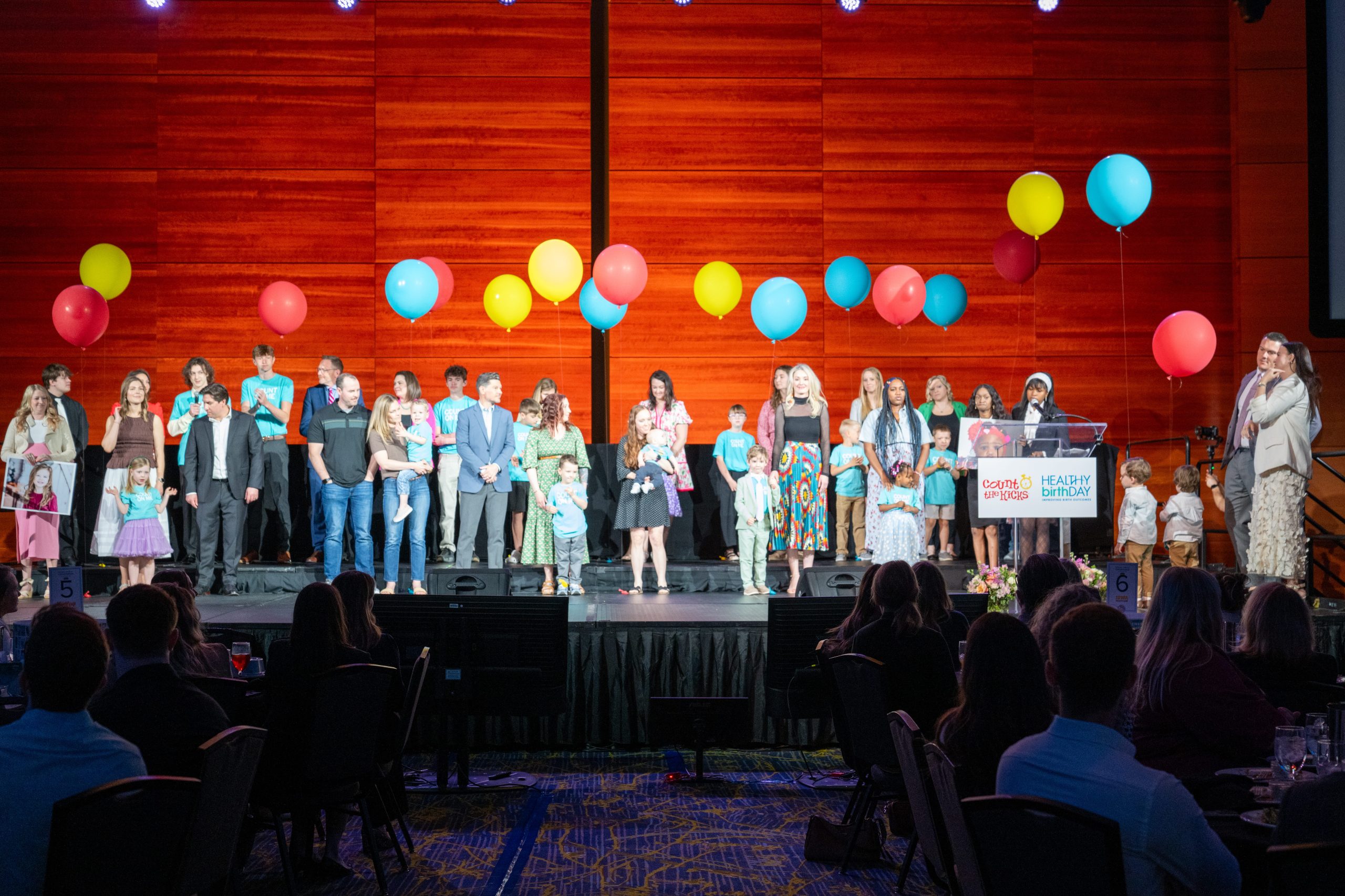 A group of people stands on a stage with colorful balloons against a wooden backdrop. They are dressed formally, conveying a celebratory atmosphere.